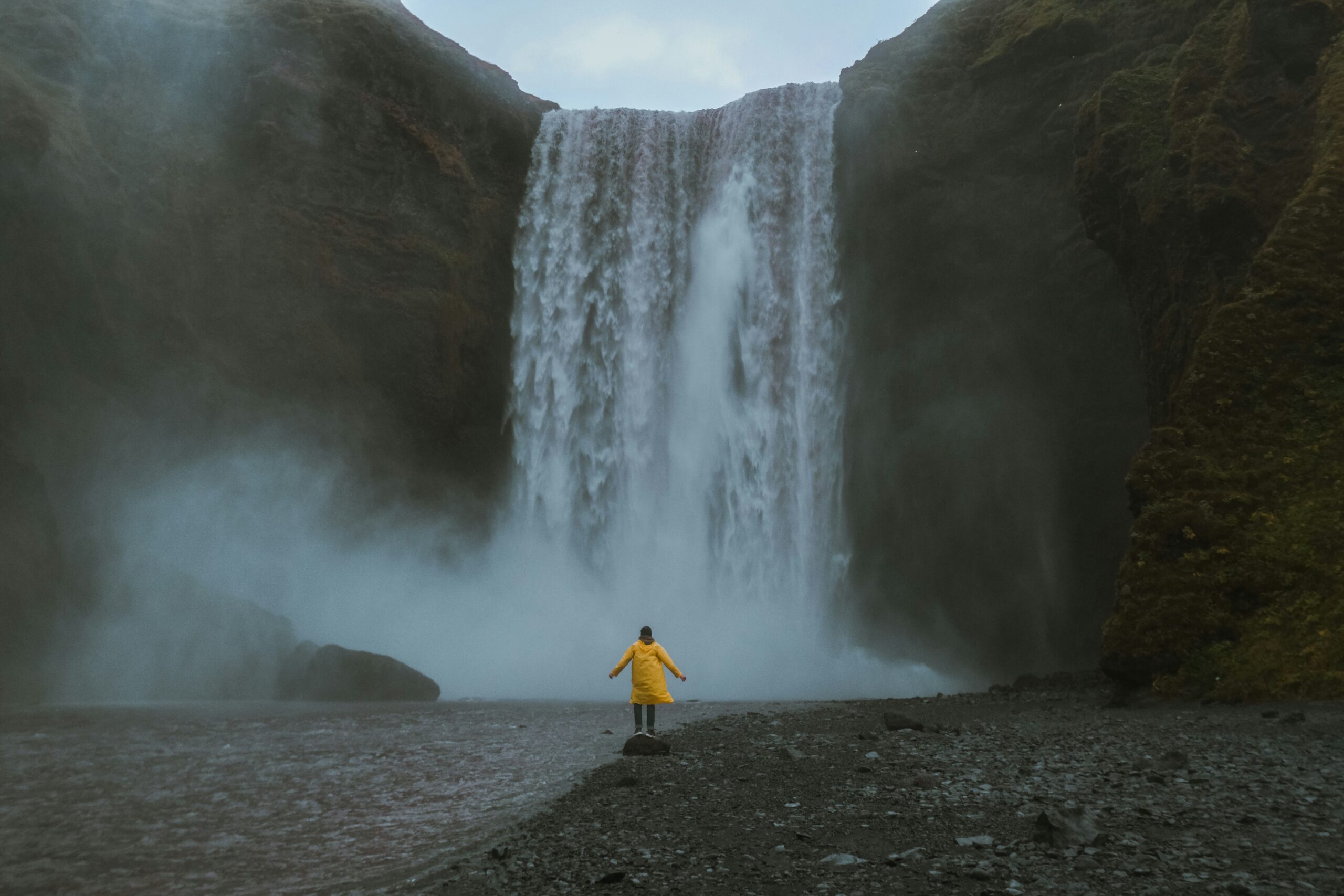 A smiling traveler standing confidently under a waterfall mist, protected by a bright yellow hooded rain poncho.